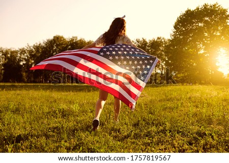 Back view. Girl with American flag runs in nature at sunset. Independence Day USA.