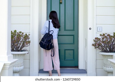 Back View Of Girl College Student Carrying Backpack Back To Home Using Key Opening Door. Young Office Lady From Work Unlock House To Enter The Room. Two Plants Beside Front Door In White Wooden House