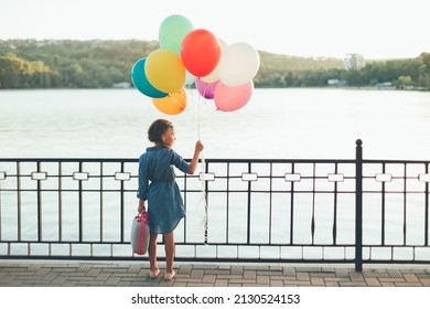Back view of girl holding colorful balloons and suitcase, in the city park enjoying wonderful view with lake, green trees and blue sky, imagining she travels. Imagination concept.