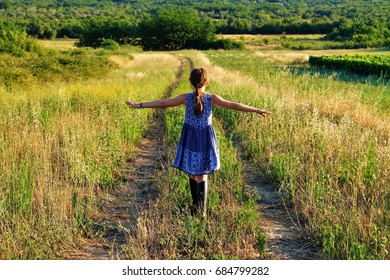 back view of girl with a plait with wide spread arms going along a path in a meadow/happy child with a dress and boots at the country/happiness, carefreeness, freedom, of luck, happy childhood