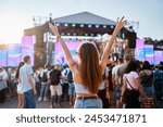 Back view of girl with raised arms, enjoying live music at sunset beach festival. Friends dance, celebrate in crowd, summer vibe at coastal event. Teens in casual wear, fun at sunny seaside gig.