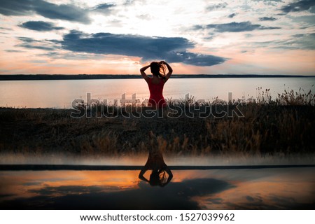 Back view of a girl in a red dress sitting on the grass near the lake, touching her hair and reflection of her shape in the water on the sunset