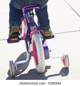 Back View Of Girl Riding Bicycle With Training Wheels.