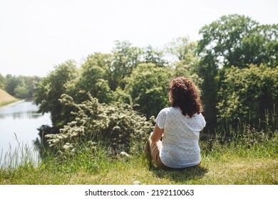 Back View Girl Traveler Sitting On Green Grass And Looking At Lake And Trees. Woman With Curly Hair. The Simple Pleasure Of Solitary Silence. Digital Detox. Copy Space For Text.