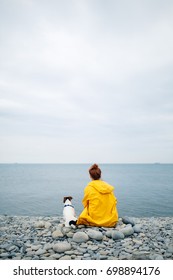 Back View Of Girl In Yellow Raincoat Sitting With Dog On Pebble Beach Looking Away At Sea.