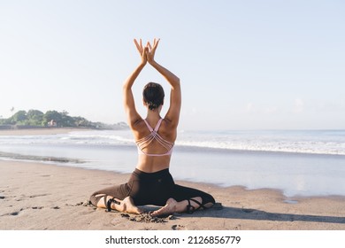 Back view of good looking female pray with raised hands recreating at coastline beach during sunny daytime, slim Caucasian woman in casual tracksuit meditate during weekend pastime at seashore
