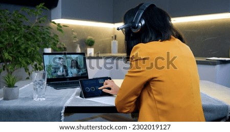 Back view of good-looking concentrated black-haired woman in headphones which listening online webinar from international colleagues and revisioning diagramms on tablet device