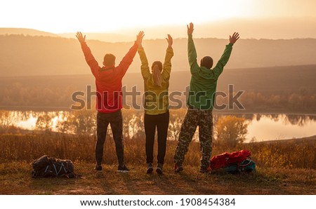 Back view of group of unrecognizable friends in activewear with backpacks raising arms and enjoying freedom while standing near lake, and resting after trekking at sunset time in autumn countryside