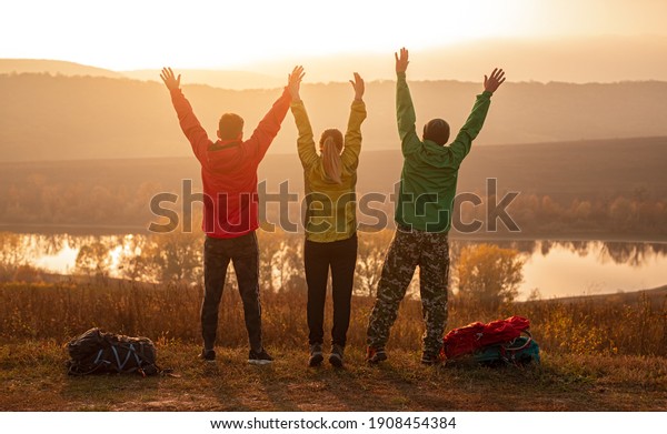 Back view of group of unrecognizable friends\
in activewear with backpacks raising arms and enjoying freedom\
while standing near lake, and resting after trekking at sunset time\
in autumn countryside