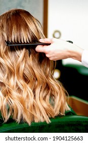 Back view of hairdresser combing wavy hair of a young blonde woman in a beauty salon