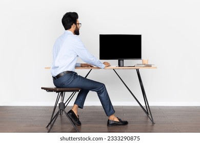 Back view of handsome young arab businessman looking at pc computer with blank black screen for mockup. Eastern man sitting at desk at home office, free space. People, technology, business concept - Powered by Shutterstock