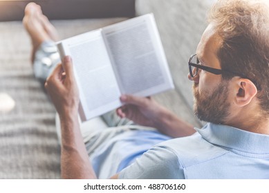 Back view of handsome young businessman in casual clothes and eyeglasses reading a book while lying on couch at home - Shutterstock ID 488706160
