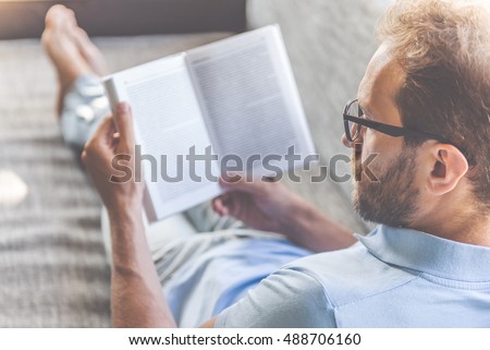 Back view of handsome young businessman in casual clothes and eyeglasses reading a book while lying on couch at home