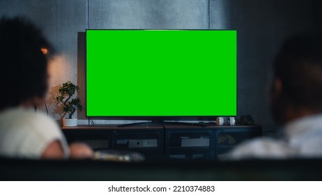 Back View Of The Happily Married Black Couple Eating Popcorn On The Sofa And Watching TV With Green Screen Mock Up Display In Loft Living Room. Out Of Focus Couple Close Up Shot At Night.