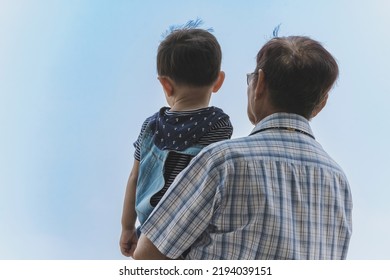 Back View Of Happiness Asian Grandfather Holding Grandson With Cloud And Sky In Background. Family Stand To Looking At Beautiful Sky Together. Joy Of Outdoor Living Together. Happy Family Relationship