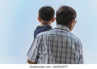 Back View Of Happiness Asian Grandfather Holding Grandson With Cloud And Sky In Background. Family Stand To Looking At Beautiful Sky Together. Joy Of Outdoor Living Together. Happy Family Relationship