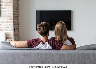 Back view of a happy couple watching tv sitting on couch at home.