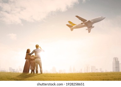 Back view of happy family embracing each other while standing at the park and looking at flying airplane in dusk sky