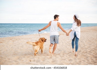 Back view of happy joyful young couple running on the beach with their dog