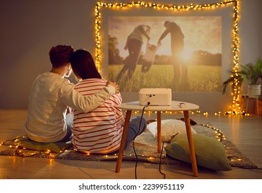 Back view happy loving couple having cozy date at home on Valentine's Day hugging on rug in living room with beautiful lights, watching movie on modern projector, looking at photos, recalling memories - Shutterstock ID 2239951119