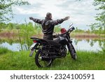 Back view of happy senior biker raising arms enjoying river landscape after riding motobike. Traveling by motorcycle. Summer road trip