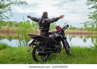 Back view of happy senior biker raising arms enjoying river landscape after riding motobike. Traveling by motorcycle. Summer road trip