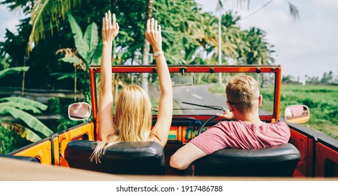 Back View Of Happy Woman With Arms Raised On Passenger Seat And Man Behind Wheel In Orange Roofless Car On Roadway In Tropical City