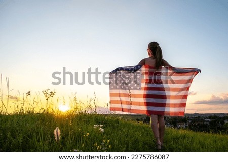 Back view of happy woman with USA national flag standing outdoors at sunset. Positive girl celebrating United States independence day. International day of democracy concept.