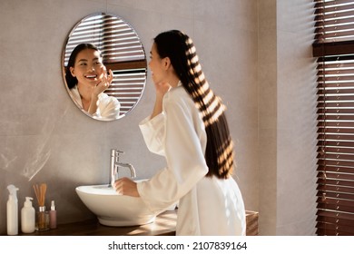 Back View Of Happy Young Asian Woman In Silk Bathrobe Applying Anti-aging Or Mosturizing Face Care Product And Smiling, Enjoying Morning Beauty Routine After Shower At Home, Copy Space