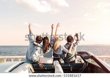 Back view of happy young friends standing with raised hands near the car