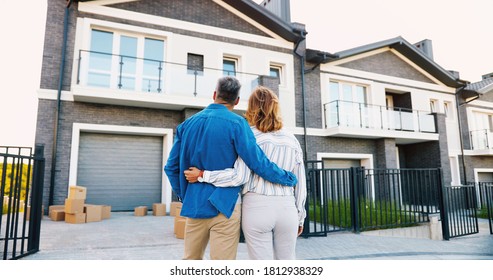 Back View Of Happy Young Just-married Couple Standing Near Their New Modern House And Hugging At Suburb And Looking On It.