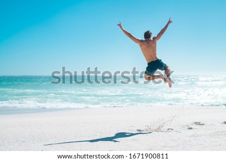 Back view of happy young man jumping with outstretched arms on the beach with copy space. Rear view of shirtless guy wearing swimwear and having fun at beach. Carefree and freedom concept on vacation.