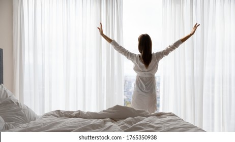 Back View Of Happy Young Woman Wake Up In The Morning In White Bright Bedroom Stretch Do Morning Exercise, Optimistic Millennial Female Awaken Look In Window Distance Meet Welcome New Day