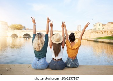 Back View Of Happy Young Women Friends Sitting With Raised Hands On The Lake In The Resort Town. Concept Of Tourism And Friendship.