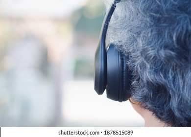 Back View Head Of A Senior Woman Wearing Wireless Headphones While Standing In A Garden. Selective Focus. Space For Text. Concept Of Aged People And Relaxation.