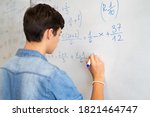 Back view of high school student solving math problem on whiteboard in classroom. Young man writing math solution on white board using marker. College guy solving math expression during lesson.