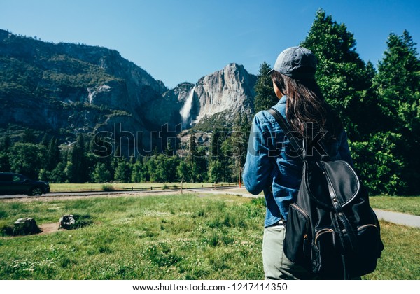 back view of hiker woman in hat walking on the grass\
sightseeing the beautiful mountain view in yosemite national park.\
young girl backpacker enjoy nature standing near path in forest on\
sunny day