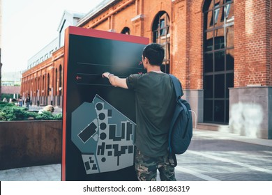 Back view of hipster guy with backpack standing in front of billboard next to old brick building and exploring scheme or map while searching for direction for walking