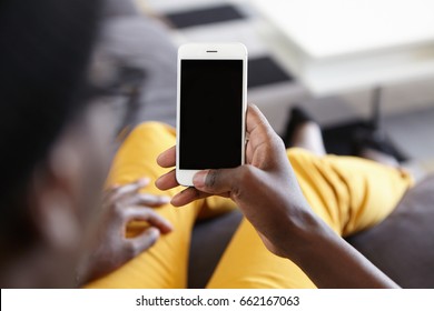 Back View Of Hipster Guy Lying At Cozy Sofa Holding Modern Smartphone With Blank Screen For Your Text Messaging And Looking Photos Online. Cropped View Of Dark-skinned Male Holding Mobile Phone