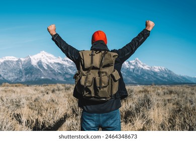 Back view of hipster guy standing with raising hands feeling excited with beautiful view of winter mountains ahead, male traveler wanderlust happy and celebrating achievement getting to destination
