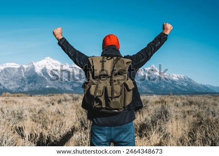 Back view of hipster guy standing with raising hands feeling excited with beautiful view of winter mountains ahead, male traveler wanderlust happy and celebrating achievement getting to destination