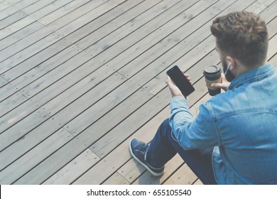 Back View. Hipster Man Is Sitting On Bench Outside, Drinking Coffee And Using Smartphone. Guy In Denim Shirt Is Sitting, Holding Phone And Cup Of Coffee, Looking At Phone Screen. Man Chatting Online.