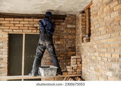 Back view of a housebuilder standing on a scaffolding and building a brick wall.