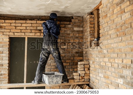 Back view of a housebuilder standing on a scaffolding and building a brick wall.