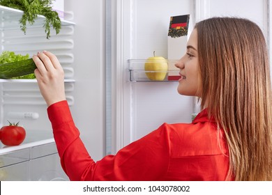 Back view of housewife wears red blouse, stands in front of opened refrigerator, chooses cucumber and tomatoes for making fresh salad, has happy expression. People, eating and nurtrition concept