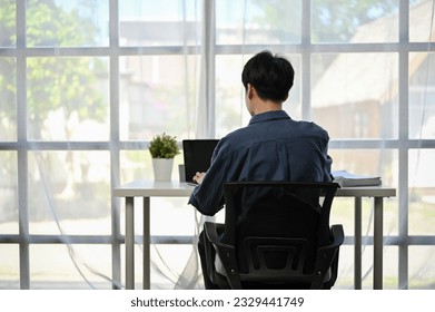 Back view image of a focused Asian businessman working on his work on his laptop computer at his desk by the window in a modern office. - Powered by Shutterstock