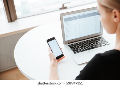 Back View Image Of Young Lady Worker Sitting In Office While Using Laptop Computer And Cellphone. Looking At Phone.