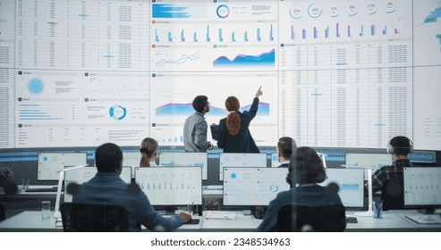 Back View Of Indian Data Analyst And Hispanic Manager Discussing Reports On Digital Screen in Monitoring Room. Male and Female Experts Talking, Employees Working On Computers In Consulting Company