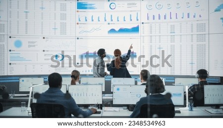 Back View Of Indian Data Analyst And Hispanic Manager Discussing Reports On Digital Screen in Monitoring Room. Male and Female Experts Talking, Employees Working On Computers In Consulting Company