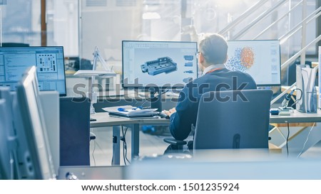 Back View of Industrial Engineer Working on Desktop Computer in Bright Office. Screens Show IDE / CAD Software, Implementation of Machine Learning, Neural Networking and Cloud Computing